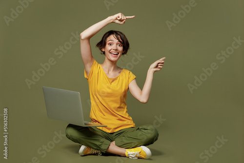 Full body young happy woman she 20s wear yellow t-shirt sitting hold use work on laptop pc computer point index finger aside on workspace area mock up isolated on plain olive green khaki background.