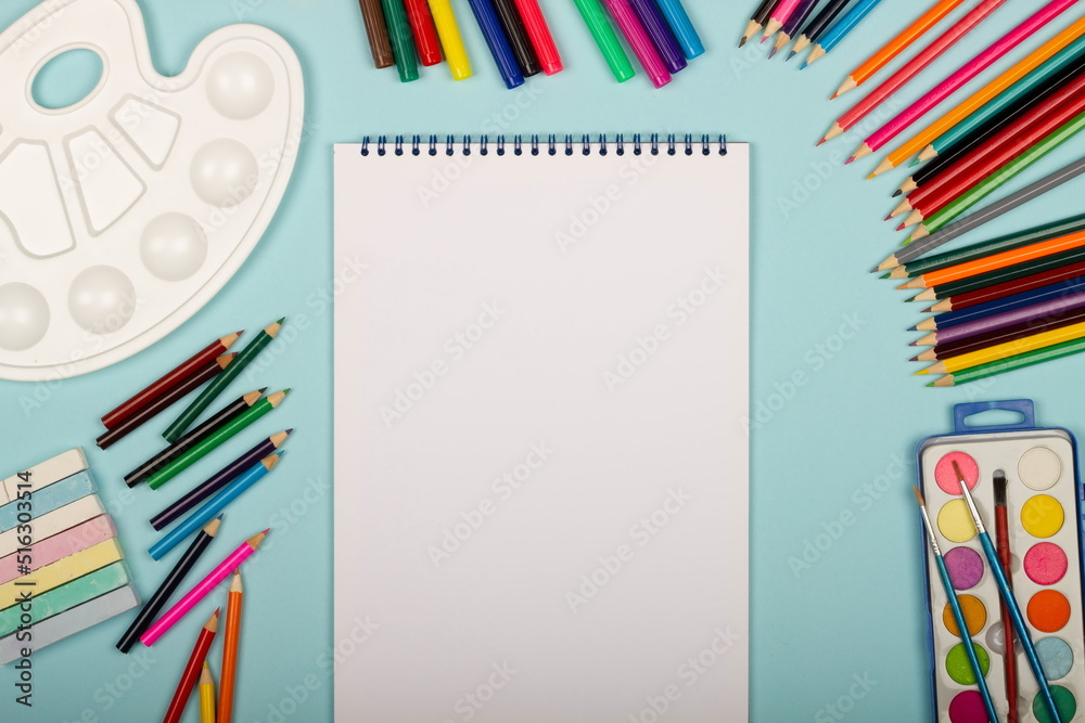 Art supplies for school painting and a blank sheet of paper on a vertical sketchbook on a blue background with copy space for text. Colorful pencils, markers, paints, crayons. Top view. Back to school Art supplies for school painting and a blank sheet of paper on a vertical sketchbook on a blue background with copy space for text. Colorful pencils, markers, paints, crayons. Top view. Back to school
