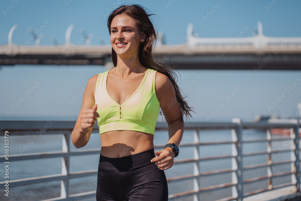 Outdoor shoot of young brunette tanned woman running among embankment, smiling broadly with happy face expression and fluttering hair. Gorgeous hispanic girl in sportswear against bay and large bridge