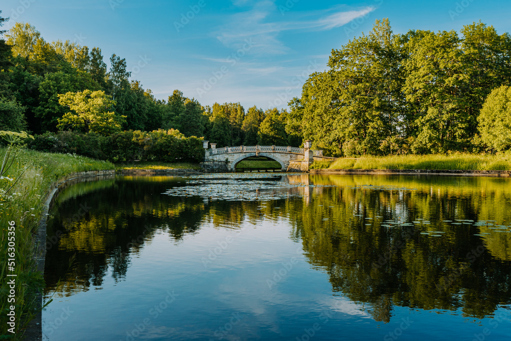 St. Petersburg, Russia, 06.22.22. View of the vintage Viscontiev Bridge and Slavyanka River in the Pavlovsk Palace and Park Complex on a summer sunny day. Trees are reflected in the water. St. Petersburg, Russia, 06.22.22. View of the vintage Viscontiev Bridge and Slavyanka River in the Pavlovsk Palace and Park Complex on a summer sunny day. Trees are reflected in the water.