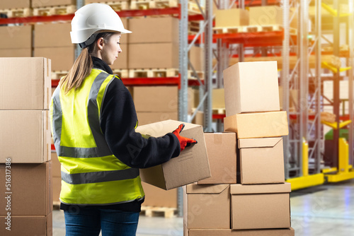 Businesswoman in warehouse. Woman with boxes at logistics center. Businesswoman in reflective vest and safety helmet. Girl in distribution center. Storage shelving blurred. Businesswoman fulfillment