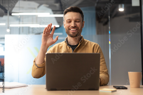 Cheerful Businessman Video Calling Using Laptop Waving Hand In Office