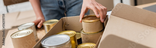 cropped view of african american volunteer packing canned food in box in charity storehouse, banner.