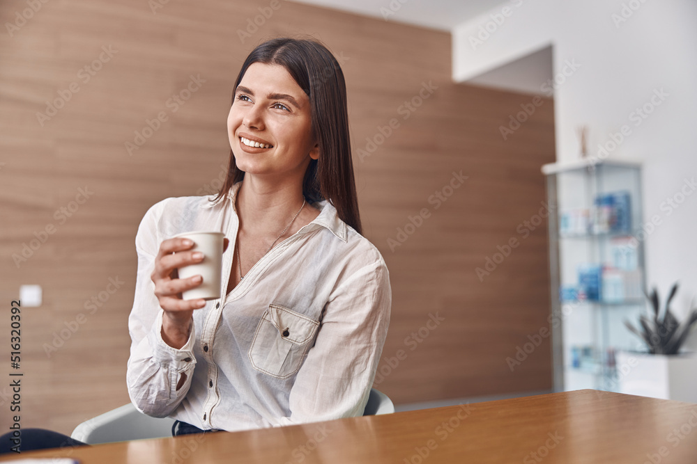 Joyful caucasian lady is drinking a coffee from a white paper cup while sitting on reception Joyful caucasian lady is drinking a coffee from a white paper cup while sitting on reception