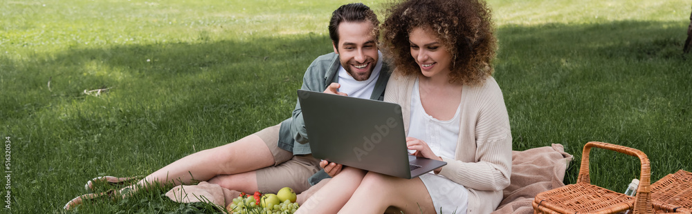 happy woman using laptop while sitting on blanket with boyfriend during picnic, banner.