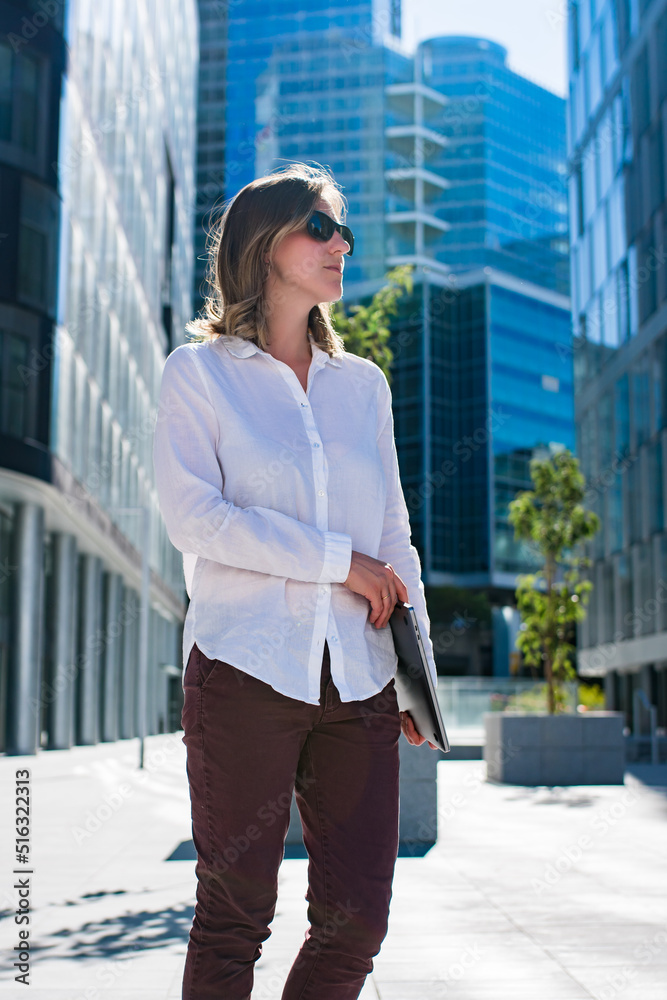 Young woman with laptop walking outdoors in business district. Woman commuting in office casual style. Portrait of a Businesswoman standing in a city district. Young woman with laptop walking outdoors in business district. Woman commuting in office casual style. Portrait of a Businesswoman standing in a city district.