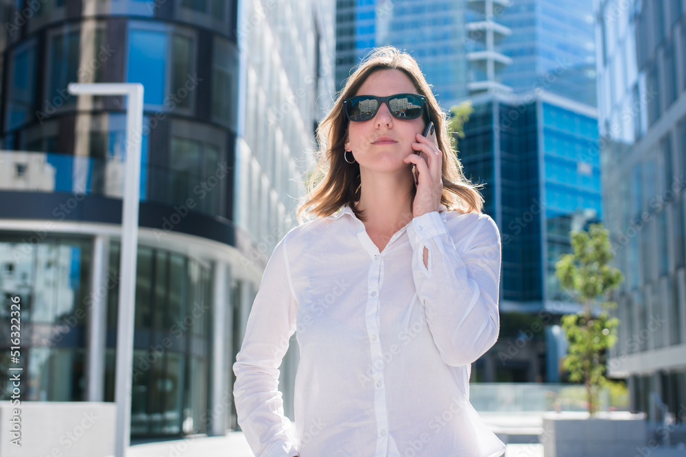 Young business woman in sunglasses stands near office centre and talks on smart phone. Business woman uses a mobile phone for work, calls. Business Woman With Phone Near Office. Young business woman in sunglasses stands near office centre and talks on smart phone. Business woman uses a mobile phone for work, calls. Business Woman With Phone Near Office.