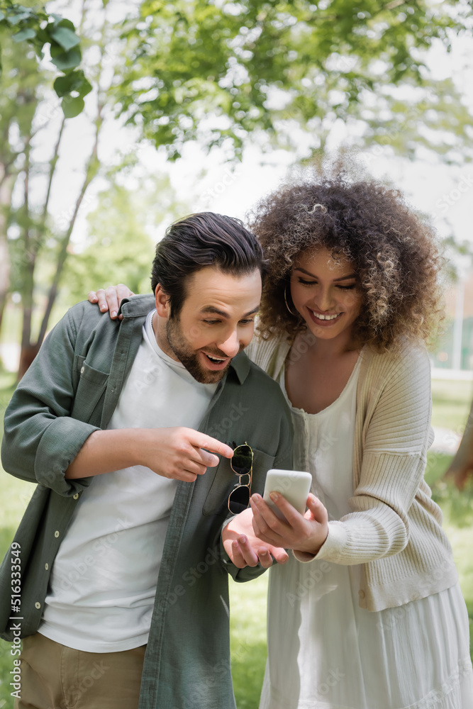 excited man pointing with finger at smartphone near curly girlfriend in green park. excited man pointing with finger at smartphone near curly girlfriend in green park.