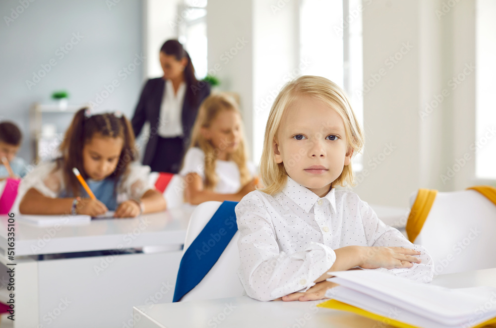 First grader elementary school. Portrait of cute little serious boy sitting at desk during lesson in school classroom. Handsome Caucasian blond kid boy sitting straight and looking at camera. First grader elementary school. Portrait of cute little serious boy sitting at desk during lesson in school classroom. Handsome Caucasian blond kid boy sitting straight and looking at camera.