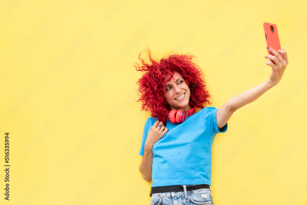 portrait of a smiling latin woman with red afro hair taking a selfie on a yellow background, wearing a blue t-shirt and red headphones around her neck.