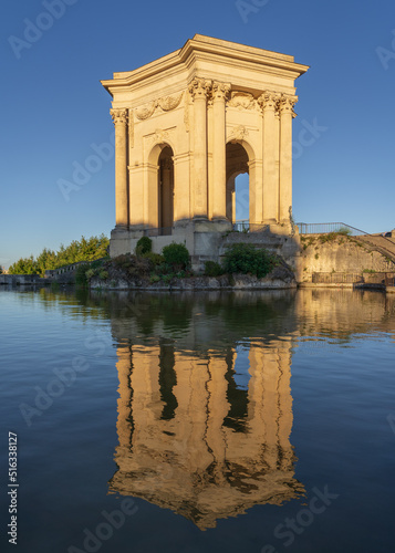 Scenic summer morning side view of historic ancient water tower stone building with reflection in water pool in landmark Promenade du Peyrou garden, Montpellier, France