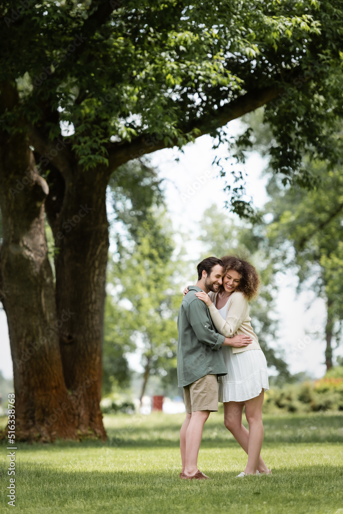 full length of cheerful couple in summer clothes hugging in park.