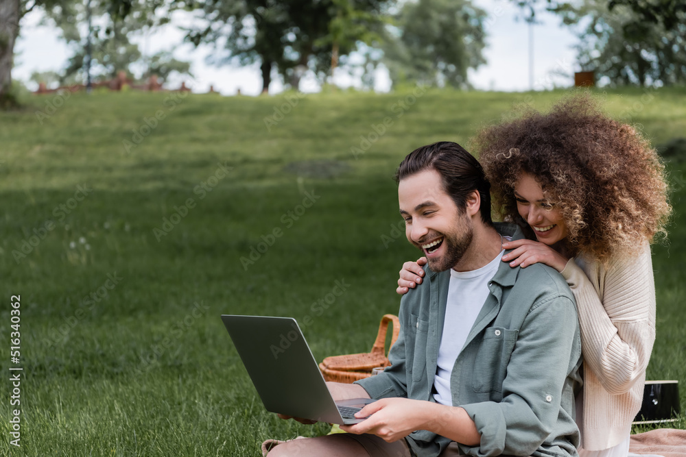 amazed man using laptop near happy woman during picnic in park. amazed man using laptop near happy woman during picnic in park.
