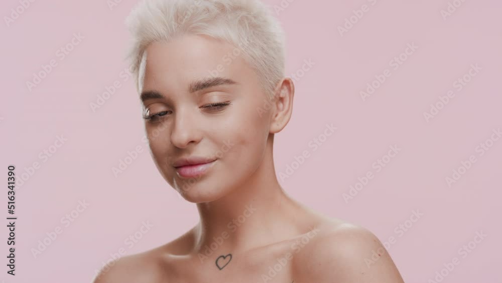 Good-looking young slim blonde Caucasian woman with naked shoulders turns to the camera smiling on pale pink background | Skin care concept