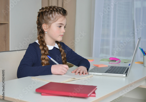 Back to school concept. Cute girl in school uniform having video conference from home using laptop and writing in notebook.

Kid looking at computer screen and taking notes.