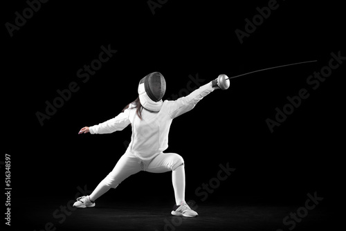 Energetic female fencer in white fencing costume and mask in action, motion isolated on dark background. Sport, youth, activity, skills, achievements, goal.