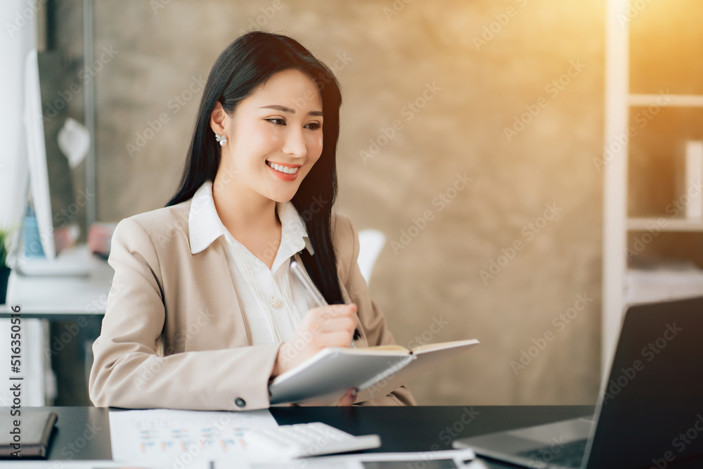Portrait of attractive woman sitting at office desk, writing on notebook and looking at laptop screen Portrait of attractive woman sitting at office desk, writing on notebook and looking at laptop screen