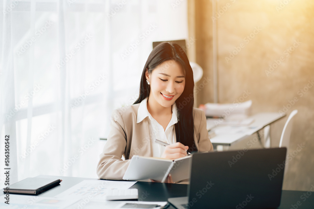 Portrait of attractive woman sitting at office desk, writing on notebook and looking at laptop screen Portrait of attractive woman sitting at office desk, writing on notebook and looking at laptop screen