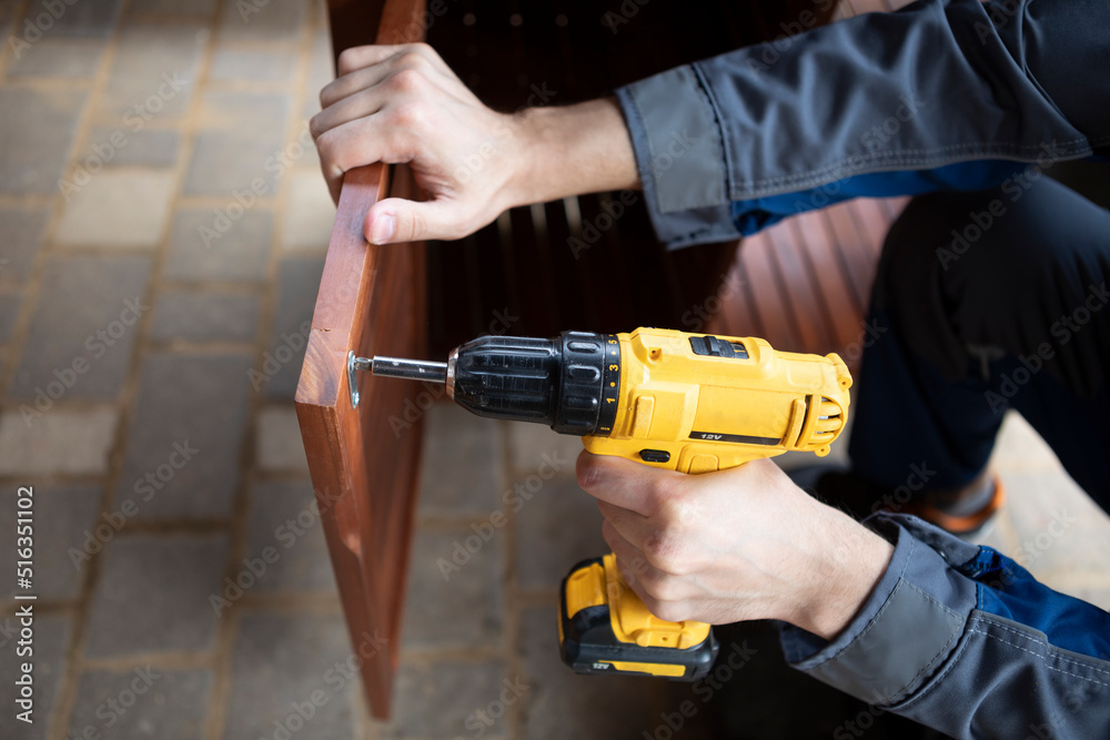 Close up hands of man holding yellow cordless screwdriver machine and assembling furniture at the yard. Fixing furniture concept