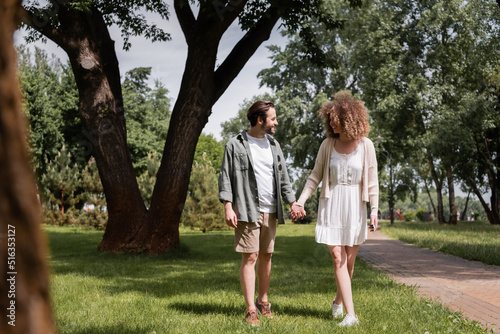 full length of happy young couple holding hands while standing on grass in summer park.