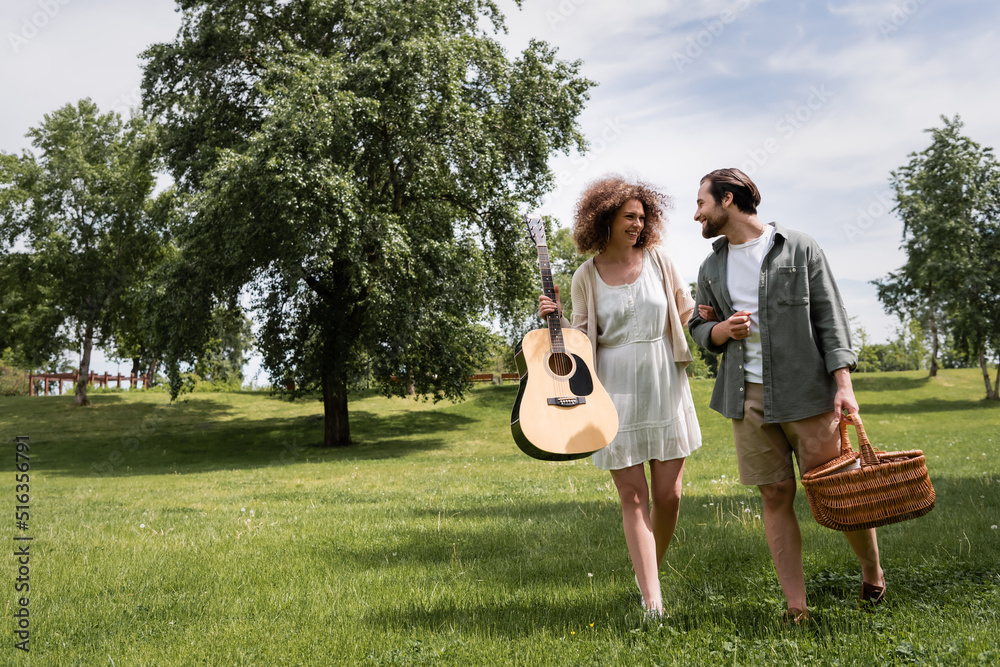 full length of smiling curly woman holding acoustic guitar near boyfriend with wicker basket walking in park.
