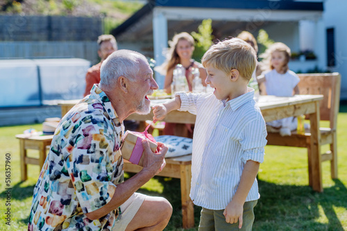 Happy little boy giving birthday present to his senior grandfather at generation family birthday party in summer garden