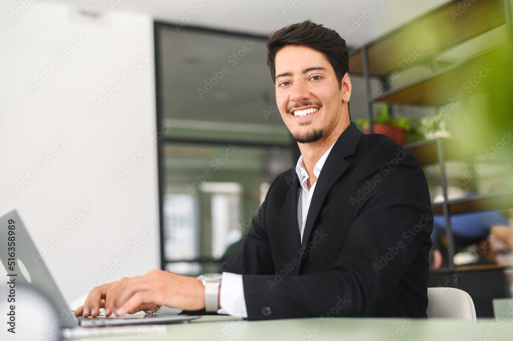 Optimistic confident dark-haired latin handsome male employee wearing formal suit using laptop for work in the office, businessman sits at the desk and looks at camera, smiles friendly