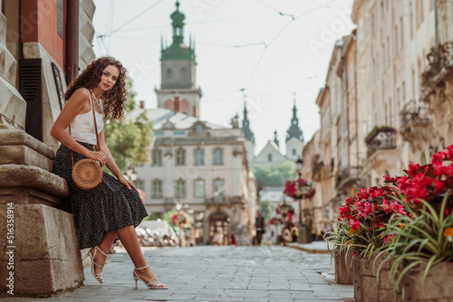 Obraz na płótnie Elegant curly brunette woman wearing trendy summer outfit with round wicker shoulder bag, white top, polka dot midi skirt, strap sandals, posing in street of European city