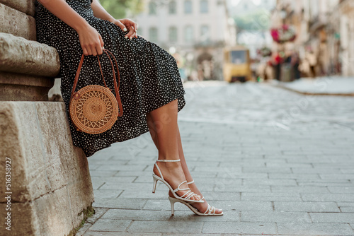 Trendy summer rotan wicker bag, white strap sandals in stylish female outfit. Woman posing in street of European city. Fashion details. Copy, empty space for text photo