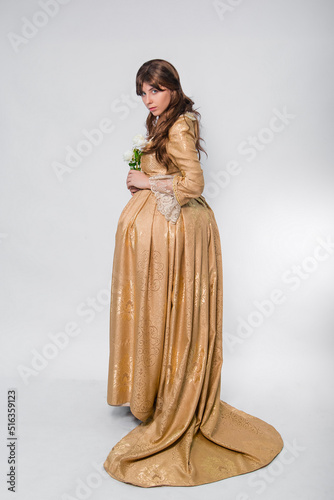 Full length portrait of a woman in a gold dress in the style of the rococo era, standing with her back forward and posing with a flower in hand isolated on a white background. 