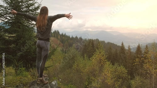 Long-haired girl on hill in green forest rejoices at setting orange sun mountains. Hands sides, hug world. Concept of amazingness world, protect nature. photo