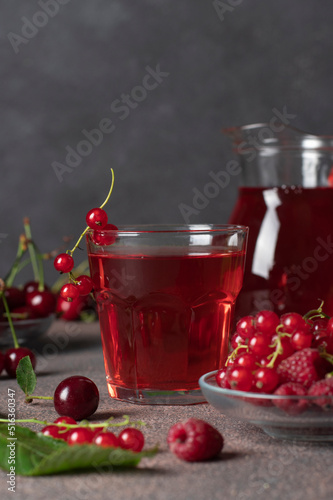 Homemade compote of red berries such as cherries  raspberries and currants in glass and jug on gray background