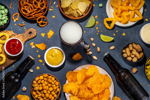 Beer with various salted snacks set. Black table background with traditional party snacks, beer bottles and glasses, with chips, onion rings, salted nuts, crisps and sauces top view copy space