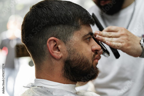 Barber guy gives a haircut to a bearded man sitting in a chair in a barbershop