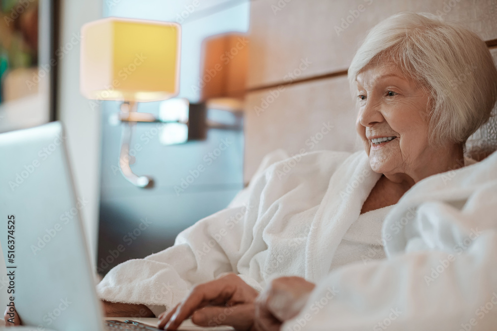 Fototapeta premium Short-haired senior woman in white robe spending time on internet and looking involved