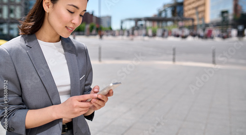 Young Asian business woman wearing suit using smartphone standing on city street outdoors. Korean lady user professional entrepreneur holding cell phone, online mobile technology business apps concept