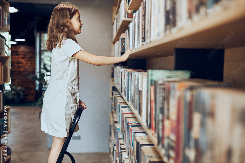 Schoolgirl looking at books on top bookshelf in school library. Smart girl selecting literature for reading. Benefits of everyday reading. Child curiosity. Back to school Schoolgirl looking at books on top bookshelf in school library. Smart girl selecting literature for reading. Benefits of everyday reading. Child curiosity. Back to school