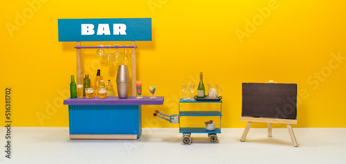 Bar service, beer drink party concept. Toy waiter trolley champagne, ice and glasses. Bar counter service area with shaker, cocktail, beer bottles spirits. Empty black chalkboard easel photo