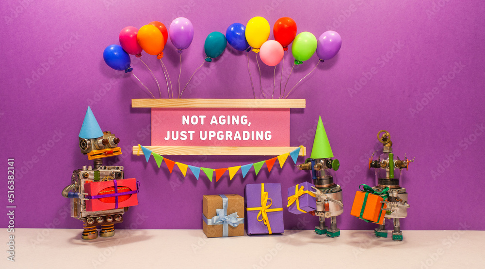 Not aging. Just upgrading. The birthday robot receives gifts from his friends. Two robots give presents cardboard boxes. banner decorated with balloons, garland of flags.