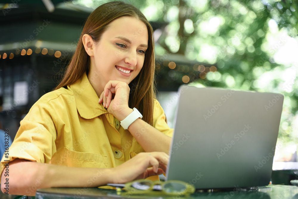 Cheerful female freelancer using laptop outdoors, enjoying remote work sitting at the table in summer cafe outdoors, smiling woman using laptop sitting in the park Cheerful female freelancer using laptop outdoors, enjoying remote work sitting at the table in summer cafe outdoors, smiling woman using laptop sitting in the park