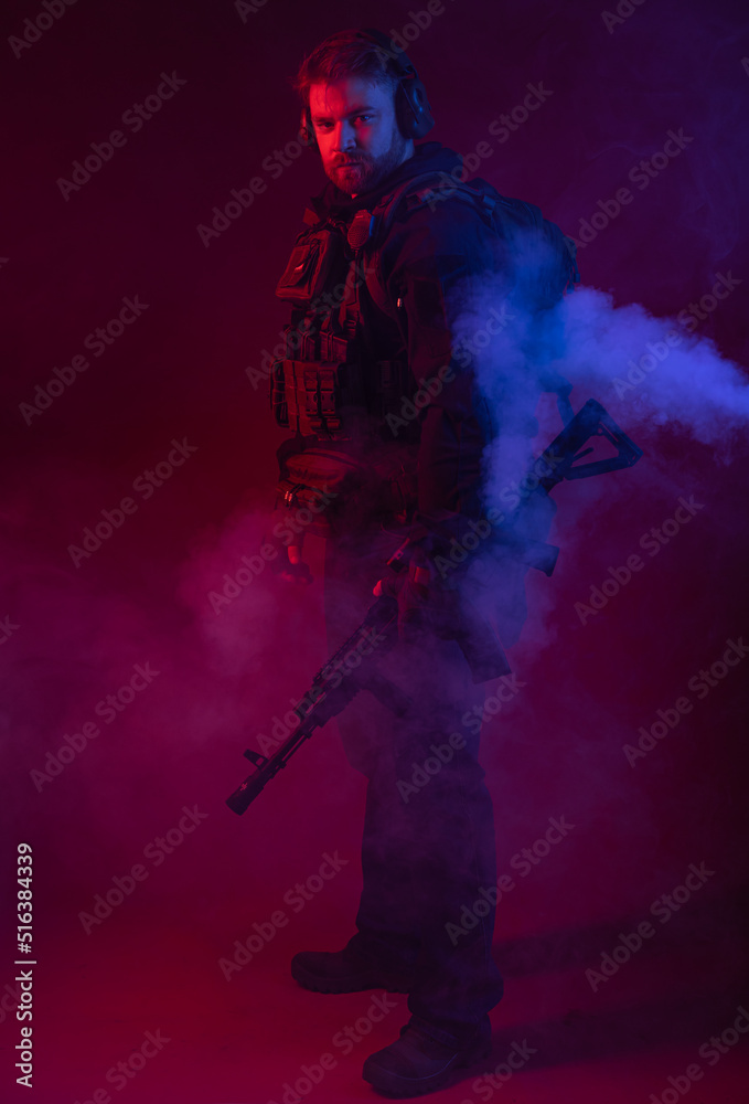 Naklejka premium soldier in full gear with weapons. a man in headphones, body armor, with a backpack and a belt. red background. colored, blue-red light. smoke around the military. explosion, chemical attack