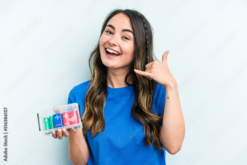 Young caucasian woman holding batteries isolated on blue background showing a mobile phone call gesture with fingers. Young caucasian woman holding batteries isolated on blue background showing a mobile phone call gesture with fingers.