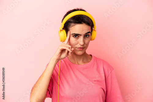 Young caucasian woman listening to music isolated on pink background pointing temple with finger, thinking, focused on a task. © Asier