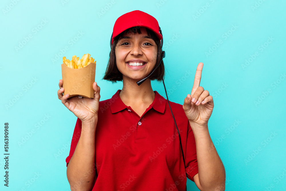 Young mixed race woman fast food restaurant worker holding fries isolated on blue background showing number one with finger. Young mixed race woman fast food restaurant worker holding fries isolated on blue background showing number one with finger.