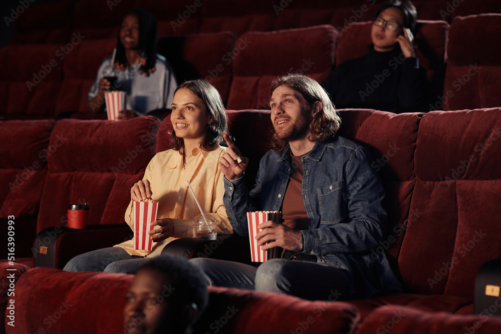 Young couple eating popcorn and watching movie together on armchairs in the cinema - obrazy, fototapety, plakaty 