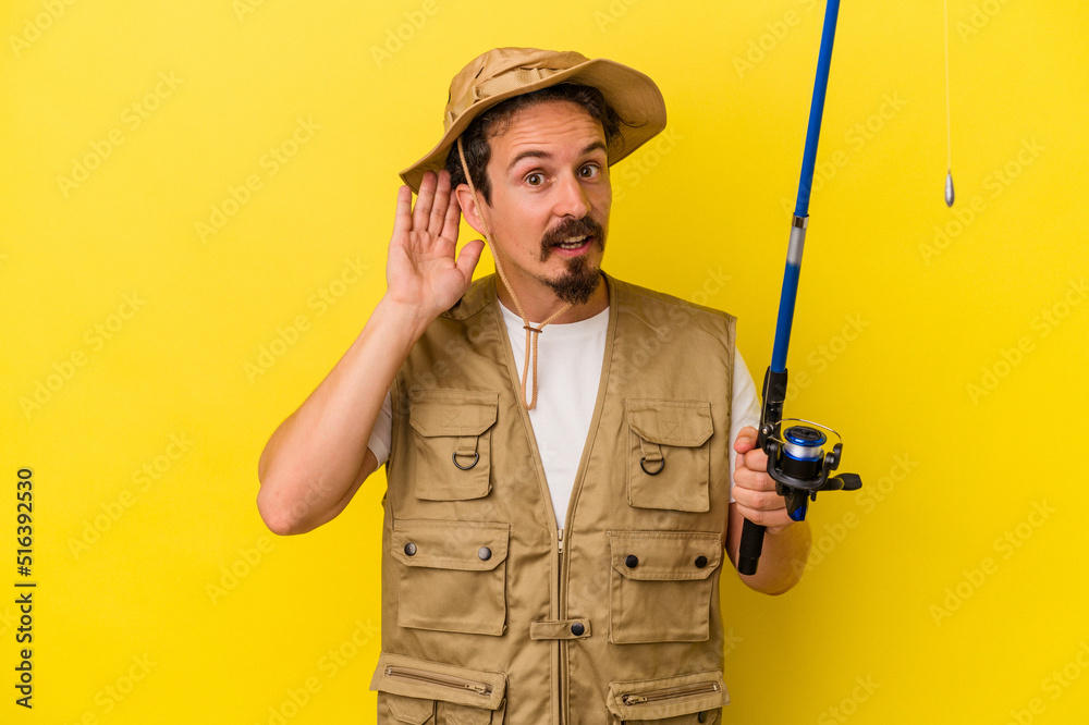 Young caucasian fisherman holding rod isolated on yellow background trying to listening a gossip. Young caucasian fisherman holding rod isolated on yellow background trying to listening a gossip.