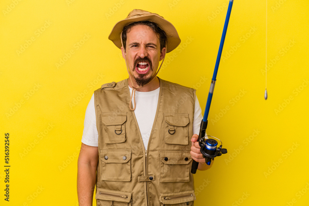 Young caucasian fisherman holding rod isolated on yellow background screaming very angry and aggressive. Young caucasian fisherman holding rod isolated on yellow background screaming very angry and aggressive.