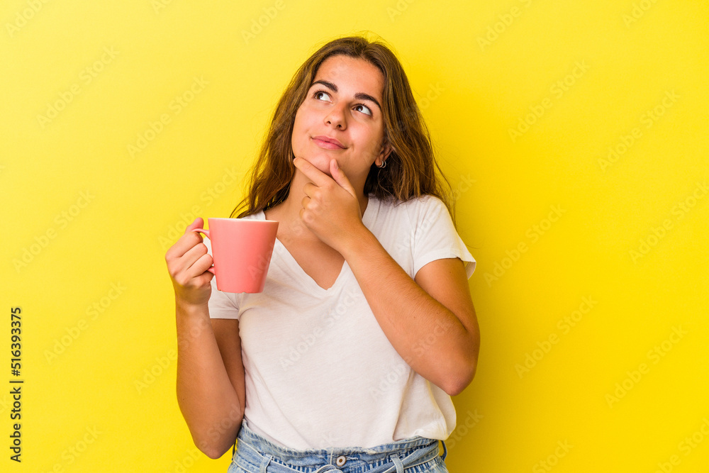 Young caucasian woman holding a mug isolated on yellow background looking sideways with doubtful and skeptical expression. Young caucasian woman holding a mug isolated on yellow background looking sideways with doubtful and skeptical expression.