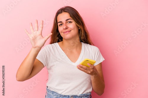 Young caucasian woman using mobile phone isolated on pink background smiling cheerful showing number five with fingers.