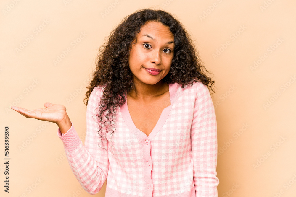 Young hispanic woman isolated on beige background doubting and shrugging shoulders in questioning gesture. Young hispanic woman isolated on beige background doubting and shrugging shoulders in questioning gesture.